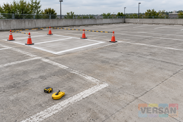 Eski beyaz çizgileri görünen beton otoparkta konilerle ayrılmış yenileme bölgesi ve ölçüm araçları
